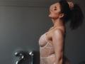Dreia vestindo uma blusa de renda branca transparente, em pé de perfil, com a mão no rabo de cavalo, olhos fechados, usando um colar e brincos, e com um sorriso discreto, mostrando seus pequenos seios e traseiro esguio..