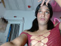 IdiiaLatina has medium brown skin and black pigtails. She wears a red, mesh, cutout top with glitter, exposing her small breasts. She makes a peace sign with her right hand, her eyes wide and looking slightly upward. A small, white dot is on her forehead. The room has a white ceiling with a red heart decoration and pink neon lights. A door with vertical slats is in the background. IdiiaLatina's expression is neutral. The image is a selfie.
