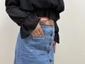 Tilliana's hand in her blue denim skirt pocket, showing pink nails, with a black sheer blouse, button-down, and brown buttons, against a plain white wall.