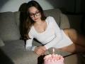 Diantrisy with glasses, white long-sleeve bodysuit, and no pants, blows out candle on pink cake, sitting on grey couch, brown hair, light skin, focused expression, dim light.