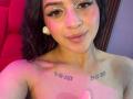 LonxEva with wavy brown hair, light skin, and eyeliner, smiling, topless, showing small tits, tattoos on chest, pink and red lighting, white earbuds, and subtle makeup.