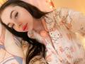 VelvetDoll with long black hair, pale skin, and pink lipstick, wearing cute pink Hello Kitty pajamas, lying in bed with a pillow. She's looking at the camera with a slight smile.