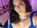 Chili has dark wavy hair, brown eyes, and a subtle smile. She's wearing a blue dress, showing tattoos on her chest and arms, with medium-sized tits.