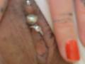 La vulve foncée et poilue de Shanna, avec un piercing en forme d'anneau doré, est touchée par un doigt portant un vernis à ongles rouge vif..