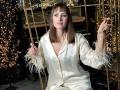 MiraMixi with brown hair in a shiny white satin pajama set, holding golden bars with white feather cuffs, smiling at the camera. Her tits are small, she's cute.