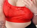 KarKutie's red lace bra, showing her tits, with a cross necklace, pale skin, yellow nails, tattoos on arms, and a pink wristband, in a tight, slightly sheer fabric.