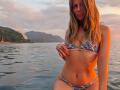 AuroraStar in a blue and white patterned bikini, standing in water, her hand on her small tits, with wavy blonde hair, pink nails, and a sexy smile.