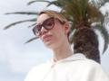MissFrenchy with short blonde hair, wearing large brown sunglasses and a white hoodie, small gold earrings, looking slightly up, with a blurred palm tree in the background.