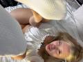 SoftAngelxKisses con capelli biondi, vestito bianco, calze bianche gonfie, sdraiata sul letto, gambe sollevate, che mostra la sua vulva, e un piccolo tatuaggio sul petto, che guarda la fotocamera..