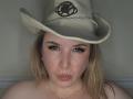 TitanicTits with blonde hair, green eyes, and thick eyelashes wearing a beige cowboy hat, pouting her pink lips, topless, showing her bare shoulders and a subtle necklace.