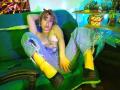 CatsitoS with pink hair, ripped jeans, yellow socks, and black boots, sitting on a green couch, topless, exposing her tits and a silver belly button ring.