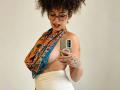 ScarlethVictoria with curly hair, glasses, and tattoos, wearing an orange patterned halter top exposing her big boobs, and white high-waisted pants, taking a selfie with a smartphone.