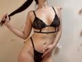 Tisia in black lace lingerie, holding a puffy brown ponytail, with a hand on her pussy, showing her tits, and a slim, toned, fair-skinned body.