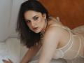 Dreia, com cabelo escuro, olhos azuis, vestindo lingerie de renda branca, um colar de pérolas e brincos, está na cama, olhando sedutivamente para a câmera, mostrando o bumbum e um leve sorriso..