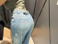 MargoFun's ass in tight blue jeans with white dragon embroidery, wearing a green shirt, standing in a store, black belt visible, black wall on the right.