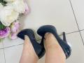Olivia Grandas glänzende schwarze High Heels an ihren Füßen, mit einem Strauß aus rosa und weißen Blumen links daneben, auf einem weißen Fliesenboden..