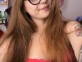 KarKutie with long brown hair, glasses, and a red lace top, showing tattoos on her arm and chest, smiling slightly. Boobs visible, nipples covered by lace.