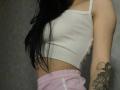 Olieri's white ribbed tank top shows her small tits, with long black hair, pink pants, and a tattooed arm against a gray wall.
