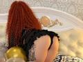 Niferttiti with red curly hair, black fishnet top, and ruffled black panties, on her knees, showing her round ass and a tattoo on her right thigh, on a white bed.