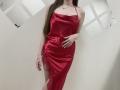 Nesie with long brown hair, wearing a shiny red satin dress with lace trim, standing, hand on her belly, visible breasts, no visible genitals, white wall in background.
