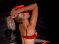 DataSley with blonde ponytail, red bra, arms raised, silver necklace, red light, dark background, showing her tits, neck, and ear, in a sexy, seductive pose.