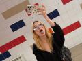 DalliaBanowski with blonde hair, glasses, and tongue out, taking a selfie in a tiled bathroom, wearing a black hoodie, holding a polka-dotted phone, and a black bag.