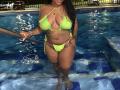 ChocolateTits with wet, dark skin, wearing a neon green bikini, holding a pool ladder, showing her big boobs and small bikini bottom, standing in a blue-tiled pool.