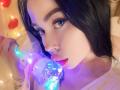 VelvetD with black hair and blue eyes, licking a glowing, translucent dildo. Her tongue touches the dildo's blue and purple lights, with a soft, white shirt on.