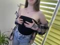 Nesie with bare boobs, hands covering black tube top, plaid shirt off shoulders, blue jeans, standing by yellow blinds, brown hair, necklace, rings, green plant in bottom left.