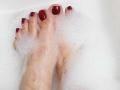 VanesaMoka's feet with dark red painted toenails, slightly submerged in white bubbly soap, showing her right and left toes. Her right toe has a small black tattoo.