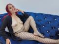 CamilaLux with red hair, smiling, wearing an open black shirt, no bra, exposing her tits, and fishnet stockings, lying on a blue tufted couch.