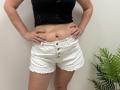 Tilliana with hands on her hips, wearing a black, cut-out, sleeveless top and white, button-up, frayed-edges shorts, showing her tan, flat stomach and pink, manicured nails.