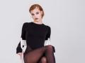 GrettaDemichelis with red hair, black long-sleeve bodysuit, and sheer black tights, sitting on a white chair, legs crossed, looking at camera with serious expression.
