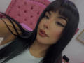 GraceClarke has long black hair, thick eyeliner, and shiny pink lips. She's wearing a light blue shirt, with a pink tufted headboard in the background.