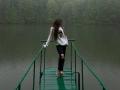 JaneKrover standing on a green metal dock, wearing a white shirt, black ripped jeans, and gray sneakers, with long brown hair covering her face.