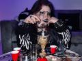 MicheMeow with dark makeup, zebra-pattern sleeves, and spiked bracelets, smoking from a gold bong, surrounded by red cups, a glass, and a red ball on a table.