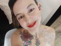 ArianaBlonde with red lipstick, brown eyes, and a smile, showing off her tattooed shoulders with colorful roses and black floral designs, wearing silver earrings, sitting on a white chair.