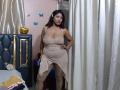 FellyLove stands in a room wearing a sheer, beige dress with a high slit, showing her big ass and big boobs. She has dark hair, a cute face, and a hand on her hip.