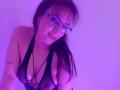 BelaDeimons, con gli occhiali, indossa un top a rete nero che mette in mostra il suo seno, ha un tatuaggio sul braccio destro ed è illuminata da una luce viola..