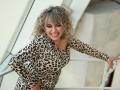 CandyLadyZ with curly blonde hair, wearing a leopard print dress, leans on a metal railing, smiling widely, showing her teeth, hands on hips, showing some cleavage.