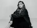 Niferttiti with wavy, medium-length hair, sitting, wearing a black satin jacket and black dress, looking at camera, legs crossed, black-and-white image, attractive, sexy, confident.