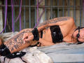 NenaBlack lies on a bed, wearing black vinyl lingerie, wrists cuffed, ball gag in mouth, tattoos on arm and thigh, purple ropes in background, textured grey sheets.