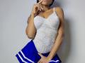 Ninah69, à la peau foncée, portant une combinaison blanche en dentelle, montrant ses seins, et une jupe bleu de cheerleader, touchant son menton et ses parties intimes avec sa main droite..