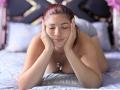 KatherineToro, with red hair, leans on a bed, hands on cheeks, eyes closed, topless, showing her tits, wearing a necklace, in a soft, purple-lit bedroom.