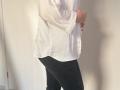 Elliance wearing a white, long-sleeve shirt with black, skinny pants. Her hand rests on her slightly curved, white-shirted belly. She stands in a softly lit room.