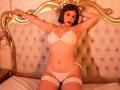 MelanyRios with pale skin, dark bob, red lipstick, wearing white lace bra and thong, arms raised, sitting on bed with golden headboard, white tufted, lace thigh-highs.
