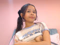 AuroraBellinii with brown hair in a ponytail, lifting a shirt to expose her small, perky tits, against a pink wall, sitting on a blue chair.