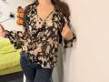 Os seios de Tilliana estão cobertos por uma blusa floral marrom, ela está vestindo jeans azuis, suas mãos estão tocando a blusa, cabelos castanhos longos, parede branca ao fundo..