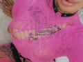 DenyChantal wearing a pink, see-through lace top with large holes, exposing tattoos on her chest and arms, and a black leather collar with a metal ring.