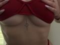 SugarrX's red ribbed bra lifts to show her tits, revealing a small, black, symmetrical tattoo just below her boobs. Her hands with long, pink nails hold the bra.